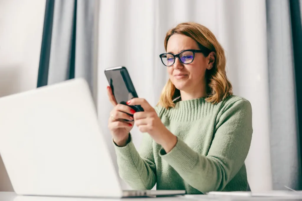Woman looking at her cellphone with a laptop on a clean desk and no papers, illustrating paperless bookkeeping habits for a calm year-end