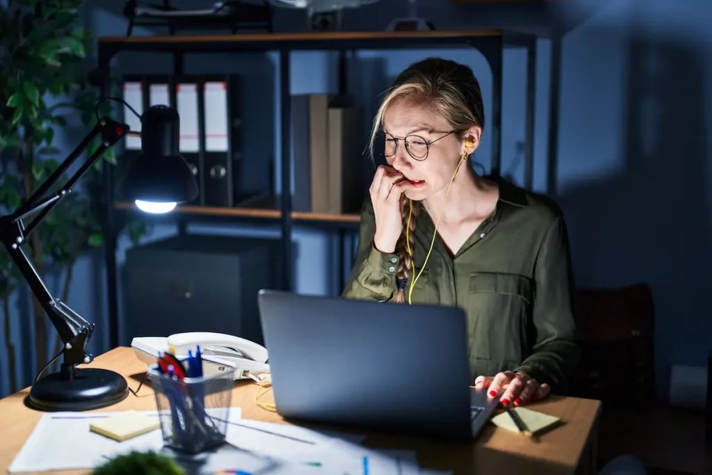 Woman sitting at her desk at night under a lamp, biting her nails while looking at her laptop screen, illustrating tax stress and the need for proactive bookkeeping habits